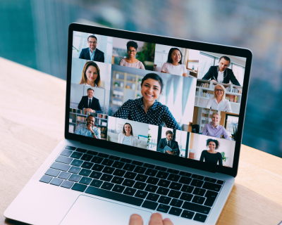Ein Schreibtisch mit einem Laptop, der ein Zoom-Meeting mit mehreren Teilnehmern zeigt, daneben ein Tablet mit einer YouTube-Oberfläche. Auf dem Tisch liegen außerdem ein Smartphone, eine Smartwatch und eine Lampe, die eine gemütliche Atmosphäre schafft.